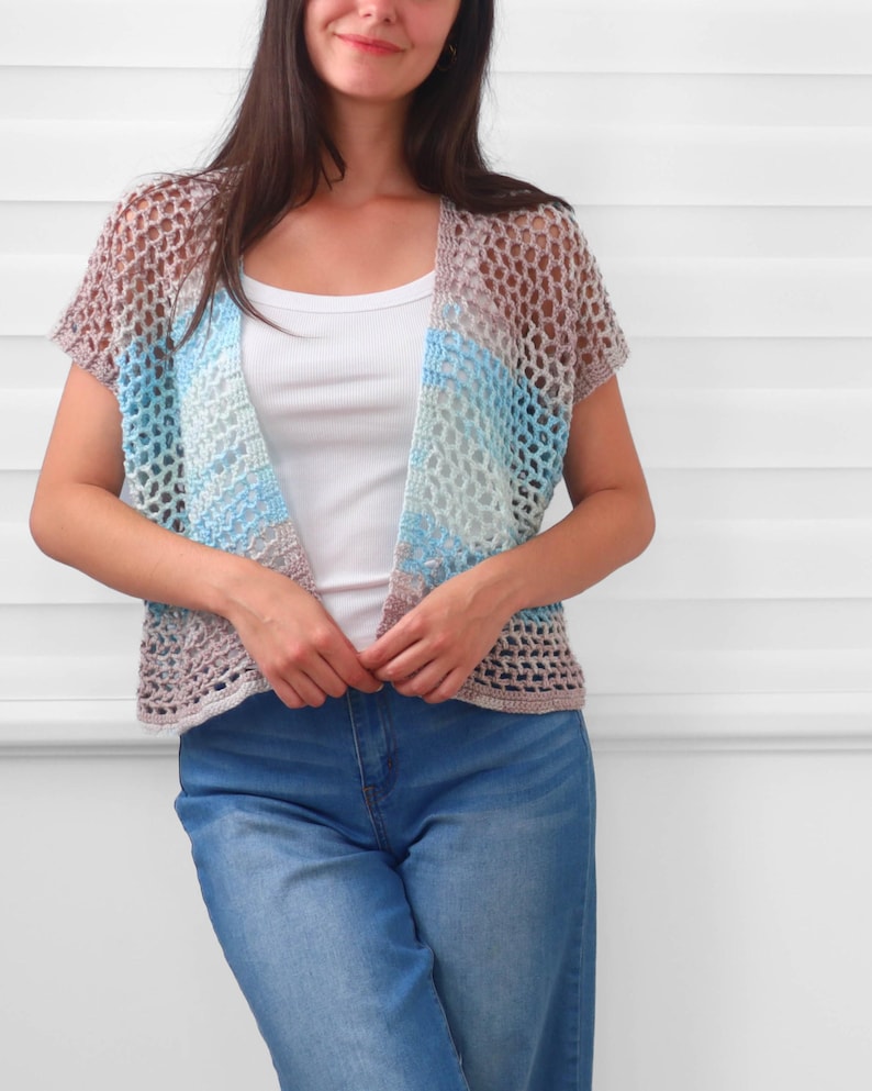 May include: A short-sleeved, open-front crochet cardigan featuring a loose, net-like stitch pattern. The garment displays a variegated color palette of soft brown, light blue, and white tones, layered over a plain white tank top and blue jeans.