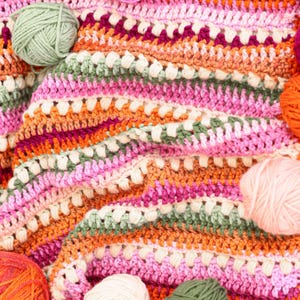Pu&ograve; includere: Una colorata coperta all'uncinetto fatta a mano con un motivo a righe nei toni del rosa, arancione, verde e bianco. Diverse palle di filo nei colori coordinati sono sparse intorno alla coperta, mostrando i materiali artigianali utilizzati.