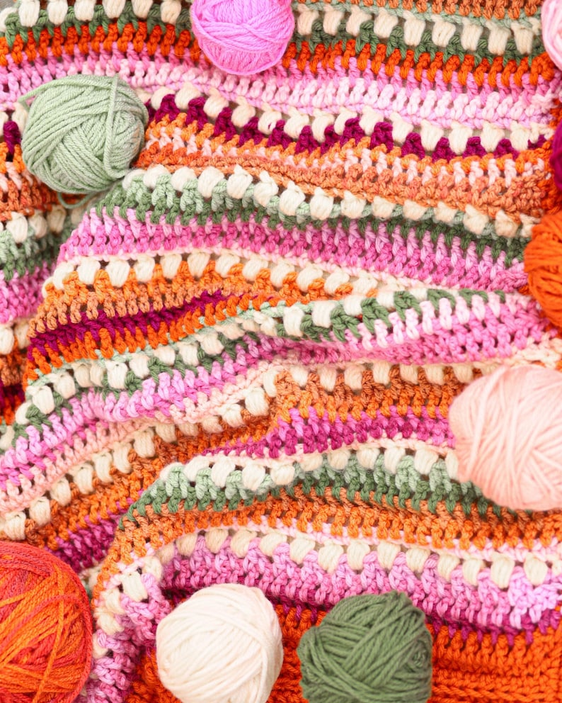 Pu&ograve; includere: Una colorata coperta all'uncinetto fatta a mano con un motivo a righe nei toni del rosa, arancione, verde e bianco. Diverse palle di filo nei colori coordinati sono sparse intorno alla coperta, mostrando i materiali artigianali utilizzati.