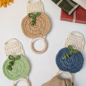 May include: Three crochet towel holders featuring round, fruit-shaped designs in tan, light green, and blue. Each piece includes a cream-colored crocheted top, a wooden ring base, and a small green crocheted leaf detail on the fruit-shaped section.