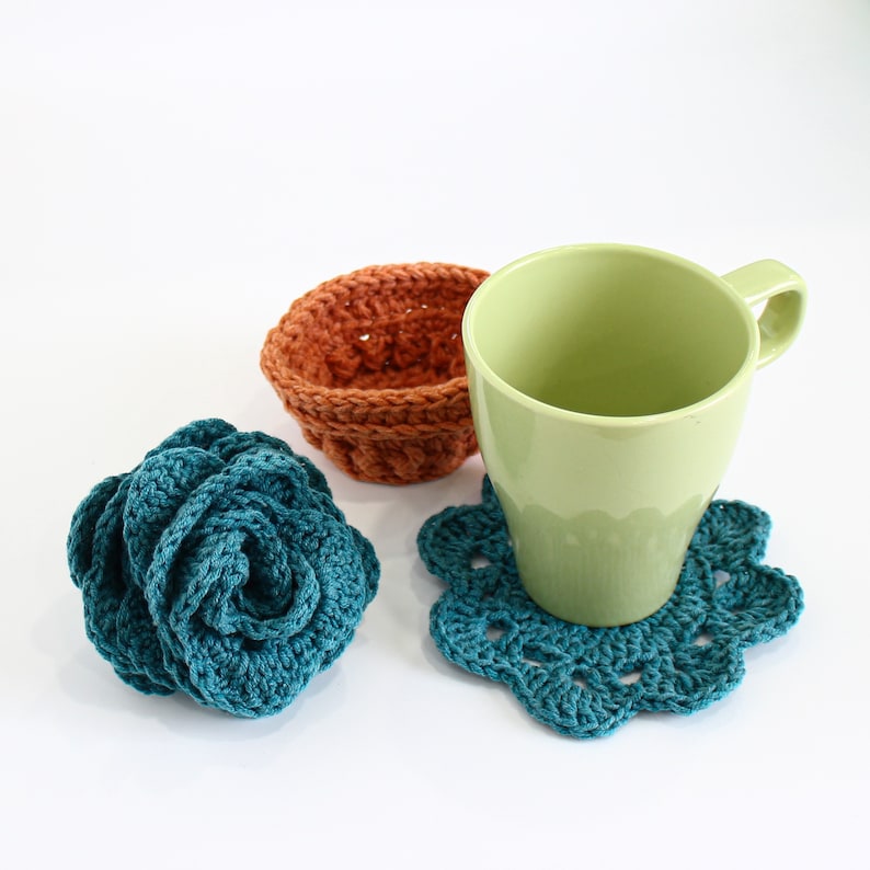 Op de afbeelding: Een set van drie gehaakte items: een teal blauwe bloem, een teal blauwe onderzetter en een bruine gehaakte kom. De onderzetter bevindt zich onder een lichtgroene keramische mok.
