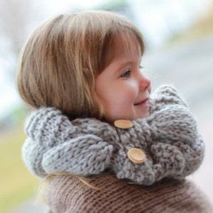 May include: A young child wearing a gray knitted cowl with wooden buttons. The cowl is thick and warm, perfect for cold weather.