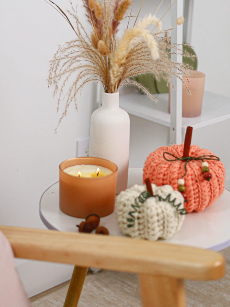 May include: A matte white ceramic vase filled with dried grasses, a frosted amber glass candle with two lit wicks, and two crocheted pumpkins in cream and coral orange, each featuring a brown stem and decorative green twine accents.