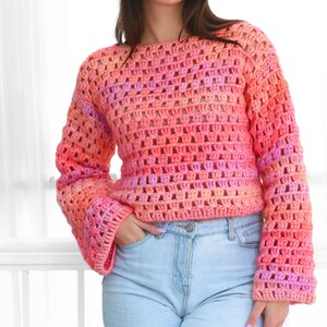 May include: A crochet sweater in shades of pink, orange, and purple. The long-sleeved top has a loose, open weave and a boat neck. The model is wearing light blue jeans. The sweater's color gradient adds a vibrant touch to the overall look.
