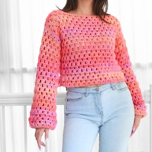May include: A crochet sweater in shades of pink and orange. The long-sleeved top has a loose, open weave design. The sweater is paired with light blue denim jeans.