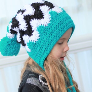 Puede incluir: Un gorro de crochet turquesa, negro y blanco con un pompón en la parte superior. El gorro tiene una larga corbata que se puede atar debajo de la barbilla.