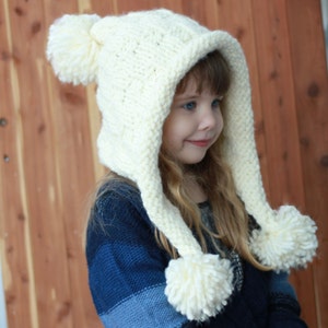 Peut inclure: Un bonnet blanc en tricot avec une longue capuche et des pompons aux extrémités. Le bonnet est porté par un enfant aux longs cheveux blonds.