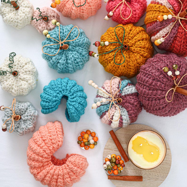 May include: A collection of crocheted pumpkins in various colors including cream, teal, orange, pink, and purple. Each pumpkin features a stem made of twine or cinnamon sticks, accented with small wooden beads and thin wire tendrils.