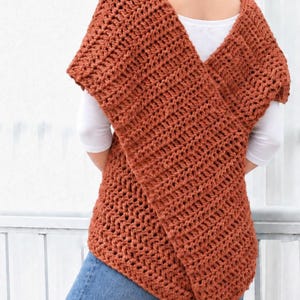 May include: A rust-colored, chunky crochet vest featuring a thick, textured knit pattern with a wide, overlapping front design. The garment is worn over a plain white short-sleeved shirt and paired with blue denim jeans, showcasing a sleeveless silhouette.