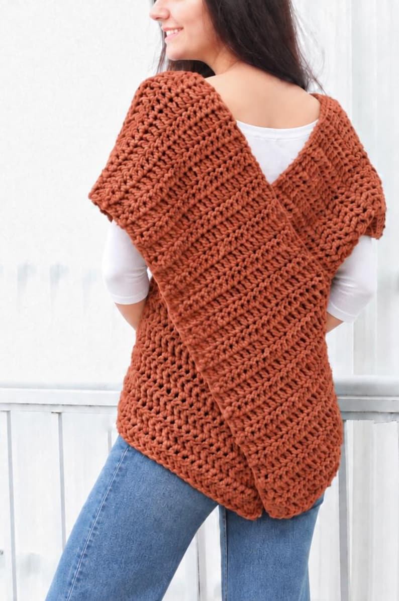 May include: A rust-colored, chunky crochet vest featuring a thick, textured knit pattern with a wide, overlapping front design. The garment is worn over a plain white short-sleeved shirt and paired with blue denim jeans, showcasing a sleeveless silhouette.