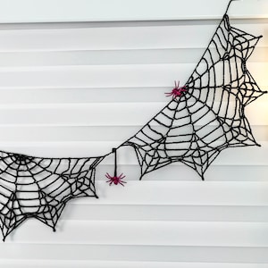 May include: A black crocheted spider web garland featuring two distinct web sections connected by a string. Two small, bright magenta plastic spiders are attached to the web structure, one resting on the upper web and one hanging below.