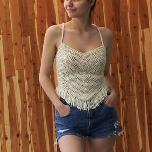 Puede incluir: Un top blanco de crochet con ribete de flecos. El top tiene cuello en V y un corte holgado. La modelo lleva el top con pantalones cortos de mezclilla.