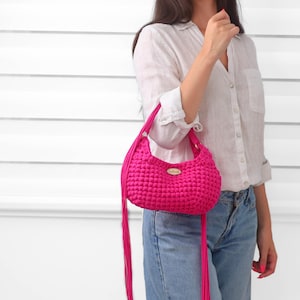 May include: A vibrant hot pink crochet handbag featuring a textured, knotted stitch pattern, a rounded silhouette, and long, thin fringe tassels hanging from the sides. A small, oval-shaped metallic brand tag is centered on the front of the bag.