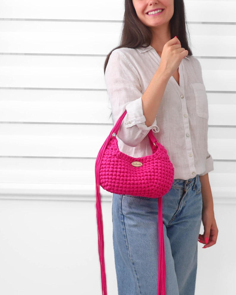 May include: A vibrant hot pink crochet handbag featuring a textured, knotted stitch pattern, a rounded silhouette, and long, thin fringe tassels hanging from the sides. A small, oval-shaped metallic brand tag is centered on the front of the bag.