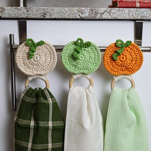 May include: Three hanging kitchen towels attached to wooden rings with crocheted pumpkin toppers in cream, light green, and orange. The towels feature a dark green plaid pattern, a solid white fabric, and a light green waffle-textured fabric.