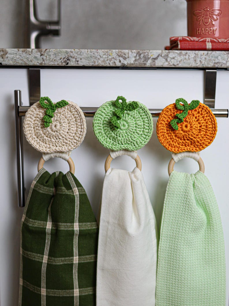 May include: Three hanging kitchen towels attached to wooden rings with crocheted pumpkin toppers in cream, light green, and orange. The towels feature a dark green plaid pattern, a solid white fabric, and a light green waffle-textured fabric.