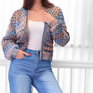 May include: A crocheted cardigan in shades of blue, tan, and brown. The open-front sweater has long sleeves and a textured stitch pattern. The person is wearing a white tank top and blue jeans.