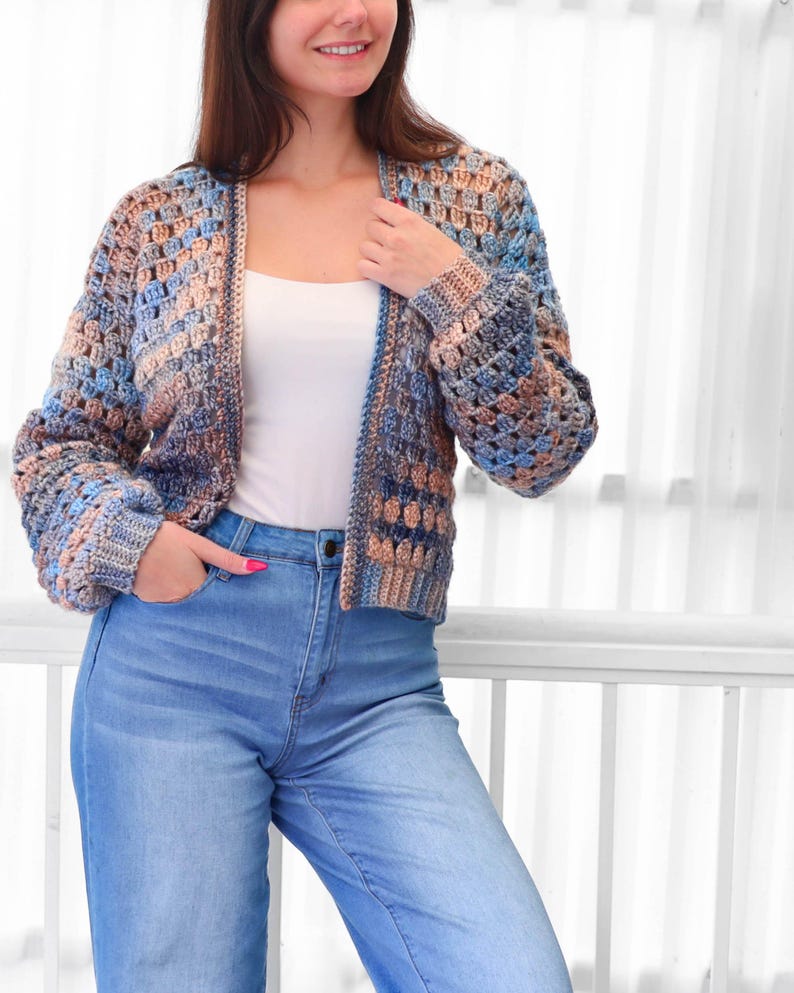 May include: A crocheted cardigan in shades of blue, tan, and brown. The open-front sweater has long sleeves and a textured stitch pattern. The person is wearing a white tank top and blue jeans.