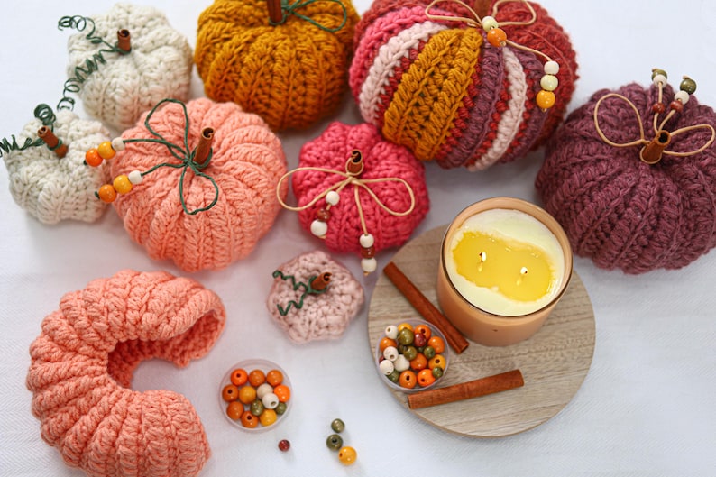May include: A collection of crocheted pumpkins in various colors including white, orange, pink, and dark purple. Each pumpkin features a cinnamon stick stem, green wire tendrils, and wooden beads. A candle in a glass jar and loose beads are included.