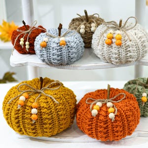 May include: A collection of handmade crochet pumpkins in various colors, including orange, yellow, and green. Each pumpkin has a twine stem, a twine bow, and decorative wooden beads. These fall-themed decorations are displayed on a white shelf.