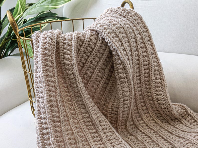 May include: A soft, beige crocheted throw blanket draped over a gold metal basket. The blanket has a textured, woven pattern.