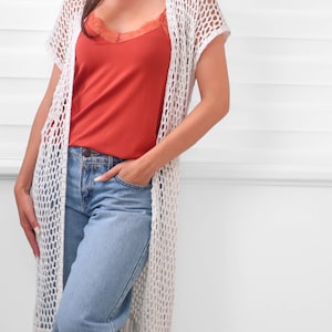 May include: A long, off-white crochet cardigan featuring an open-knit mesh pattern, short sleeves, and a fringed hem. It is worn over a rust-colored camisole with lace trim and light blue denim jeans, creating a layered look with a textured finish.