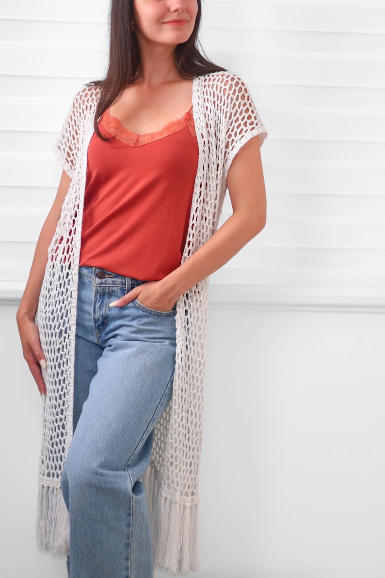 May include: A long, off-white crochet cardigan featuring an open-knit mesh pattern, short sleeves, and a fringed hem. It is worn over a rust-colored camisole with lace trim and light blue denim jeans, creating a layered look with a textured finish.