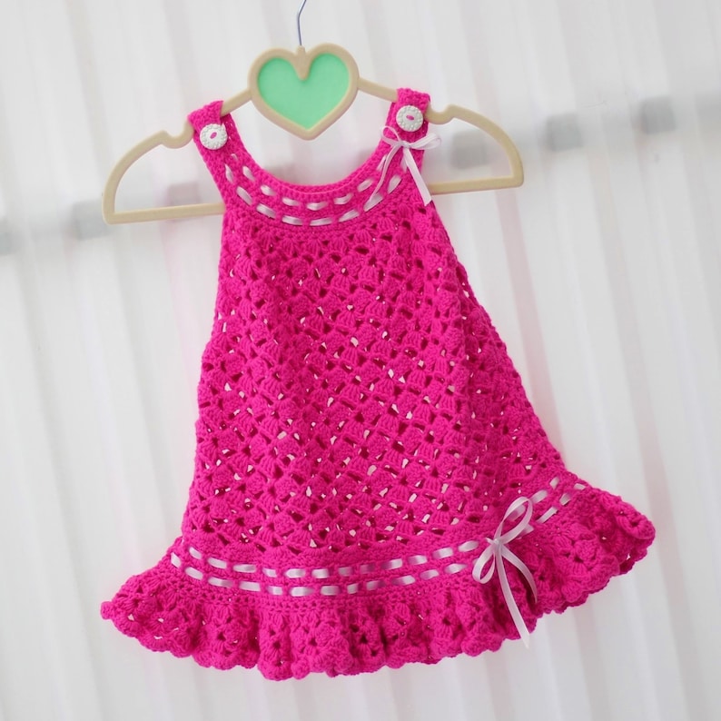 May include: A pink crocheted baby dress with white ribbon accents and white buttons. The dress has a ruffled hem and is hanging on a wooden hanger.