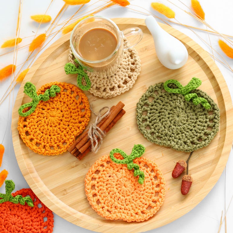 May include: Three round crochet coasters shaped like pumpkins in orange, light orange, and olive green, each with a green stem. A glass mug of coffee sits on the beige coaster, accompanied by cinnamon sticks and two small acorn decorations.