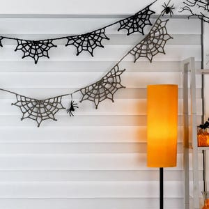 May include: A two-tiered crochet garland featuring black and grey spider web motifs connected by a thin string. Small black spider figures are attached to the webs. The garland is draped horizontally against a white horizontal blind.