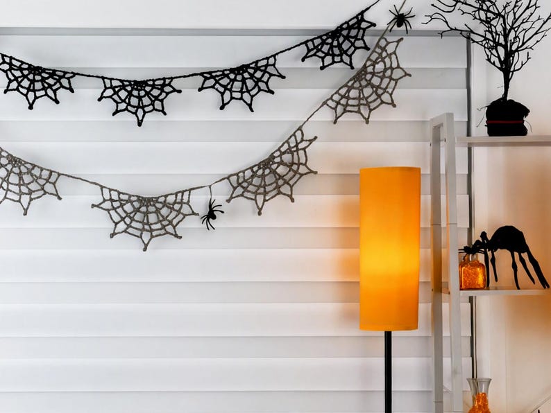 May include: A two-tiered crochet garland featuring black and grey spider web motifs connected by a thin string. Small black spider figures are attached to the webs. The garland is draped horizontally against a white horizontal blind.