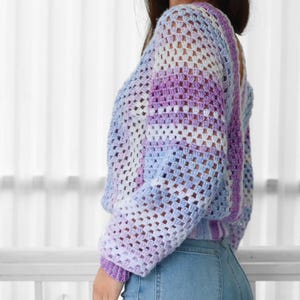 Pode incluir: Um top de croch&ecirc; em roxo claro, branco e azul, com caimento solto e mangas compridas. O top &eacute; feito com um padr&atilde;o de ponto quadrado.