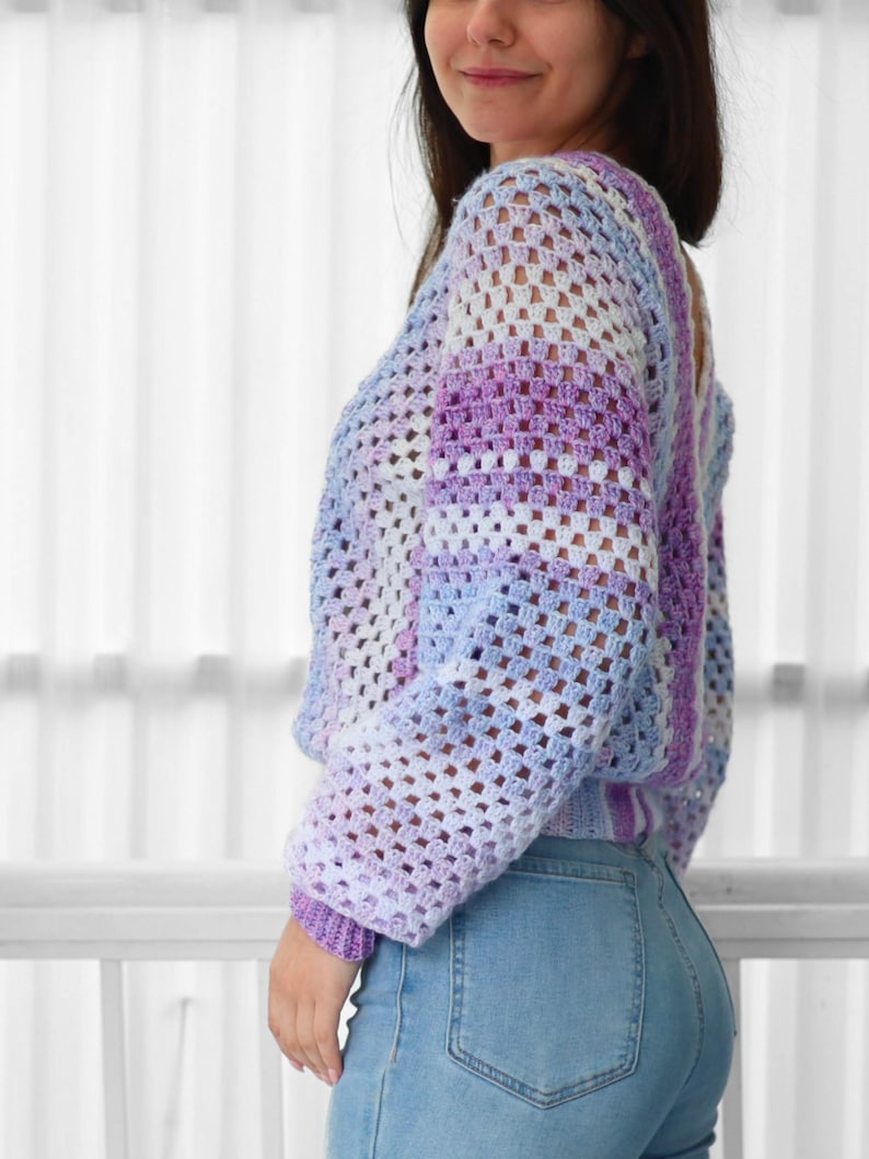 Pode incluir: Um top de croch&ecirc; em roxo claro, branco e azul, com caimento solto e mangas compridas. O top &eacute; feito com um padr&atilde;o de ponto quadrado.