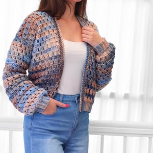 May include: A multi-colored crochet cardigan in shades of blue, tan, and brown. The open-front sweater has long sleeves and a textured, open weave design. The model is wearing blue jeans and a white tank top.
