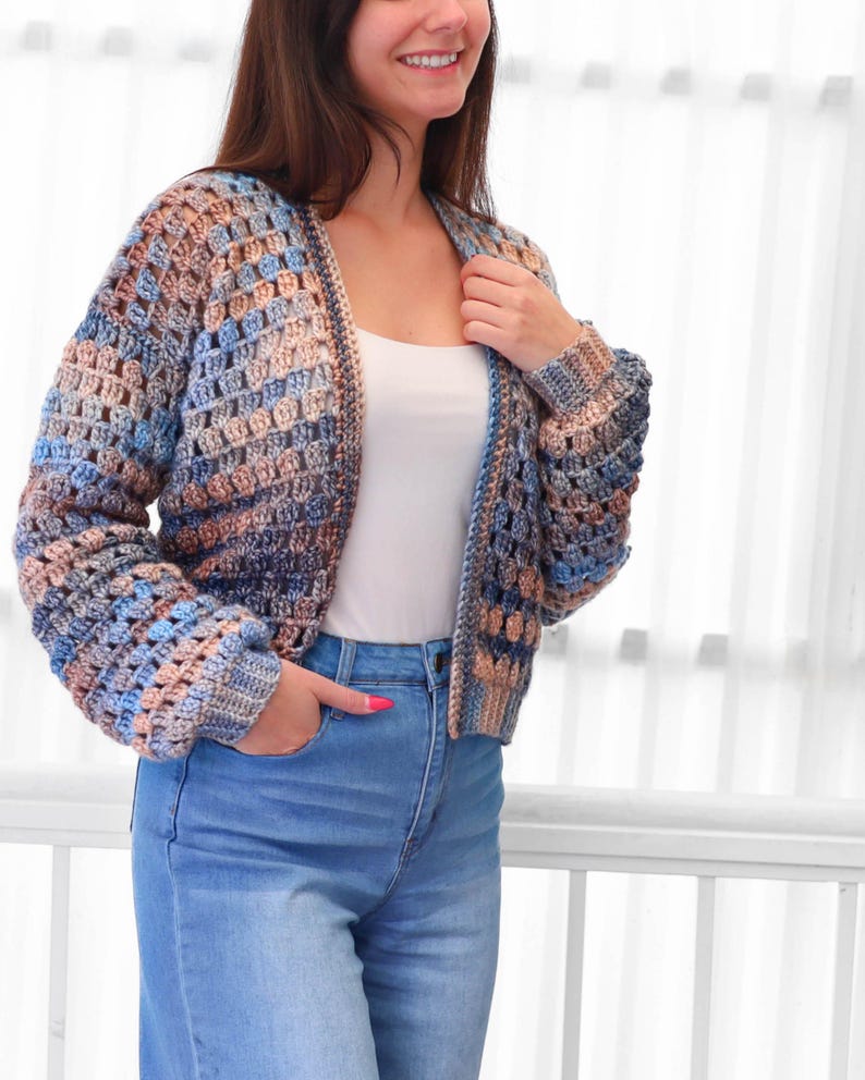 May include: A multi-colored crochet cardigan in shades of blue, tan, and brown. The open-front sweater has long sleeves and a textured, open weave design. The model is wearing blue jeans and a white tank top.