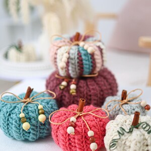 May include: A collection of six crocheted pumpkins in various colors including teal, pink, burgundy, and cream. Each pumpkin features a cinnamon stick stem, a thin twine bow, and small wooden beads. The pumpkins have a ribbed texture.
