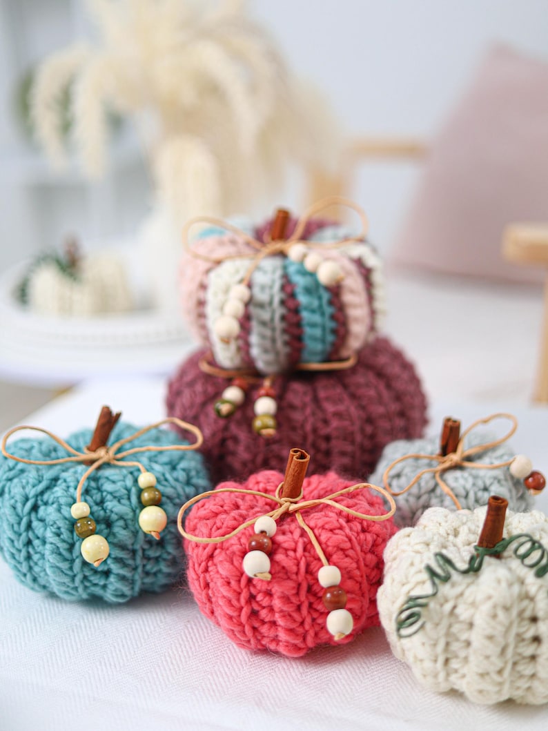 May include: A collection of six crocheted pumpkins in various colors including teal, pink, burgundy, and cream. Each pumpkin features a cinnamon stick stem, a thin twine bow, and small wooden beads. The pumpkins have a ribbed texture.