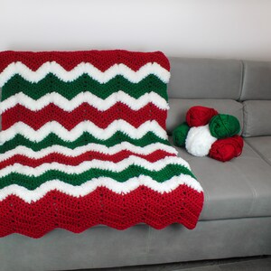 May include: A red, white, and green crocheted blanket with a chevron pattern. The blanket is on a gray couch with balls of yarn in the same colors.