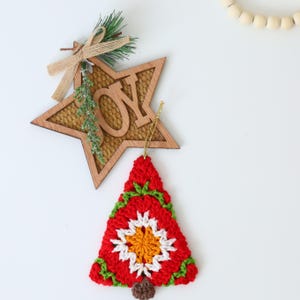 Puede incluir: Un adorno de &aacute;rbol de Navidad de ganchillo en rojo, blanco, verde y naranja con una base de fieltro marr&oacute;n. El adorno cuelga de una estrella de madera con la palabra "JOY" escrita en ella.
