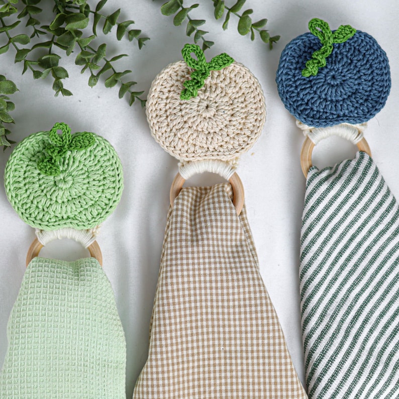 May include: Three hanging kitchen towels, each featuring a crocheted fruit topper in light green, beige, or blue, attached to a wooden ring. The towels include a light green waffle weave, a beige gingham check, and a dark green and white striped pattern.
