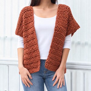 May include: A rust-colored, open-front crochet cardigan featuring a textured, ribbed stitch pattern with short sleeves. The garment is worn over a plain white t-shirt and blue denim jeans, showcasing a simple, layered look with a loose, comfortable fit.