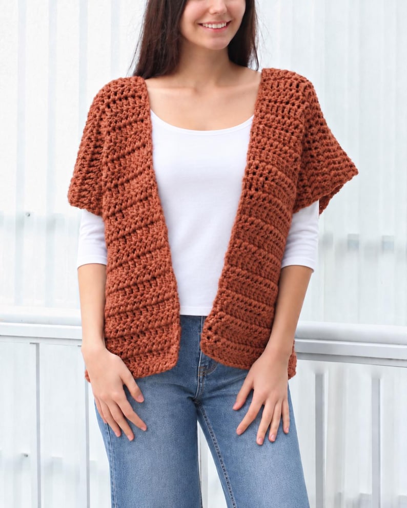 May include: A rust-colored, open-front crochet cardigan featuring a textured, ribbed stitch pattern with short sleeves. The garment is worn over a plain white t-shirt and blue denim jeans, showcasing a simple, layered look with a loose, comfortable fit.