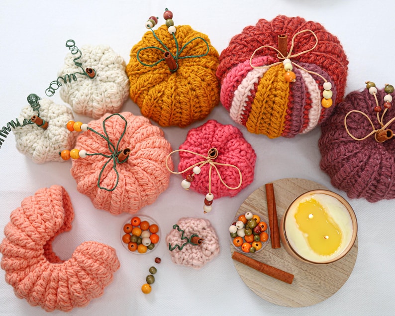 May include: A collection of crocheted pumpkins in various colors including cream, mustard yellow, pink, red, and plum. Each pumpkin features a cinnamon stick stem, green wire tendrils, and small wooden beads. A candle and loose beads are included.