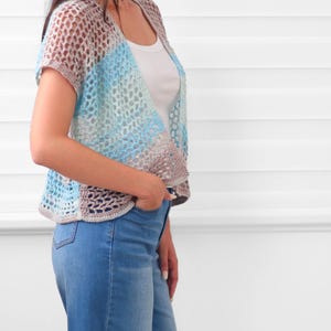 May include: A short-sleeved, open-front crochet cardigan featuring a color-blocked pattern in shades of light blue, white, and soft beige. The garment is constructed with an airy, open-stitch design and is worn over a simple white tank top and blue jeans.