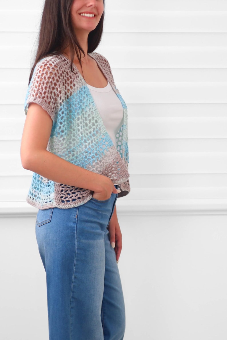 May include: A short-sleeved, open-front crochet cardigan featuring a color-blocked pattern in shades of light blue, white, and soft beige. The garment is constructed with an airy, open-stitch design and is worn over a simple white tank top and blue jeans.
