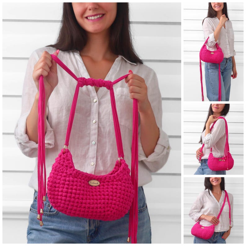 May include: A bright fuchsia crochet handbag featuring a textured, knotted stitch pattern, a long adjustable strap with dangling fringe ends, and a small oval-shaped wooden tag labeled handmade attached to the front of the bag.