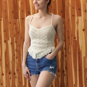 Puede incluir: Un top blanco de crochet con detalles de flecos. El top tiene un escote redondo y tirantes finos. La modelo lleva pantalones cortos de mezclilla.
