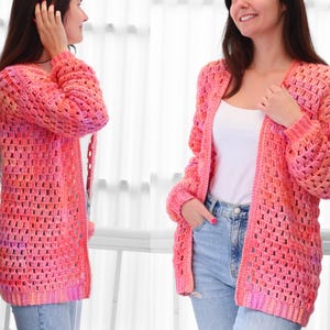 May include: A long, open-front crochet cardigan in shades of pink, orange, and purple. The handmade sweater features a textured, open weave design with long sleeves and a relaxed fit. The cardigan is worn over a white top and blue jeans.