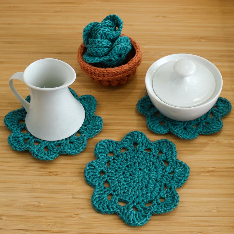 Op de afbeelding: Drie teal gebreide onderzetters met een witte keramische kan en een witte keramische suikerpot met deksel op een houten oppervlak.