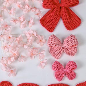 May include: Three crochet bows in varying shades of red and pink, arranged vertically. The top bow is a deep red, the middle is a soft light pink, and the bottom is a vibrant pink. A long, scalloped red crochet trim piece lies horizontally below.